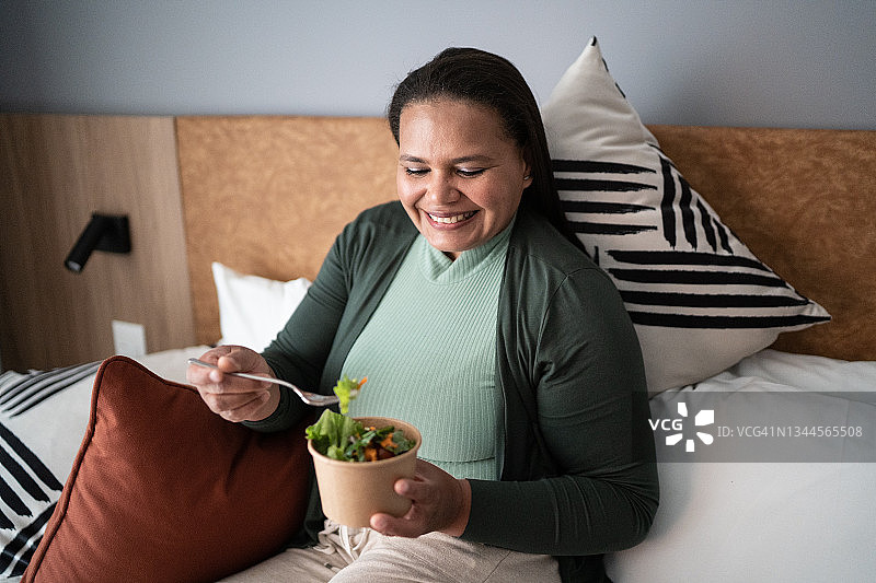 快乐的中年妇女在家里的床上吃沙拉图片素材
