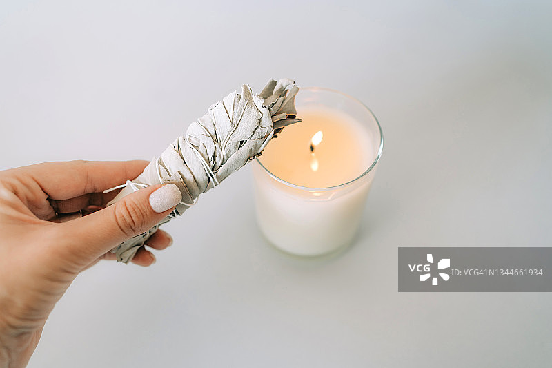 女人手持白色鼠尾草，上方是灰色背景上的燃烧蜡烛和圣木棒。用于芳香疗法和仪式的套装。顶视图图片素材