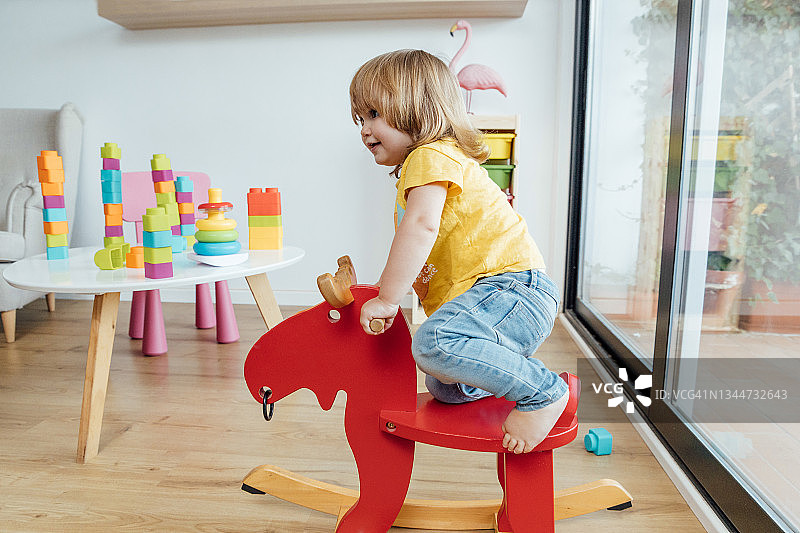 可爱的宝宝在幼儿园玩耍，骑在红色木马上，玩得很开心的侧视图图片素材