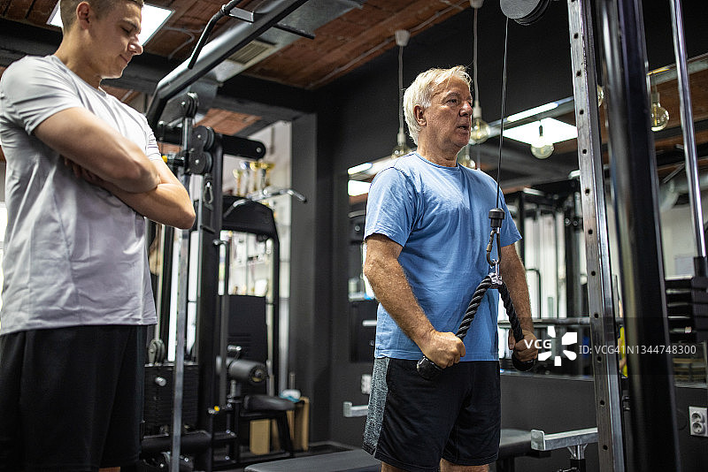 年长的男人在健身房的器械上锻炼，旁边有私人教练图片素材