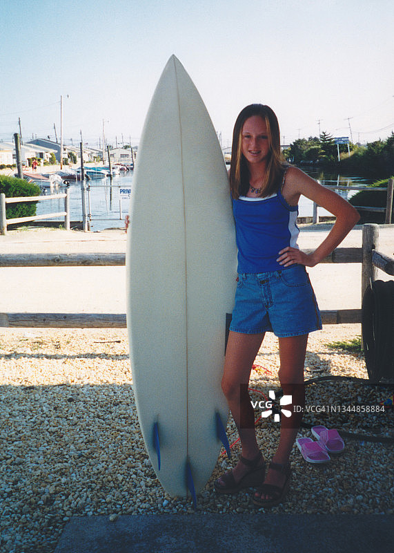 20世纪90年代少女：具有90年代冲浪女孩风格的少女在夏季海滩度假时在冲浪板旁摆姿势图片素材