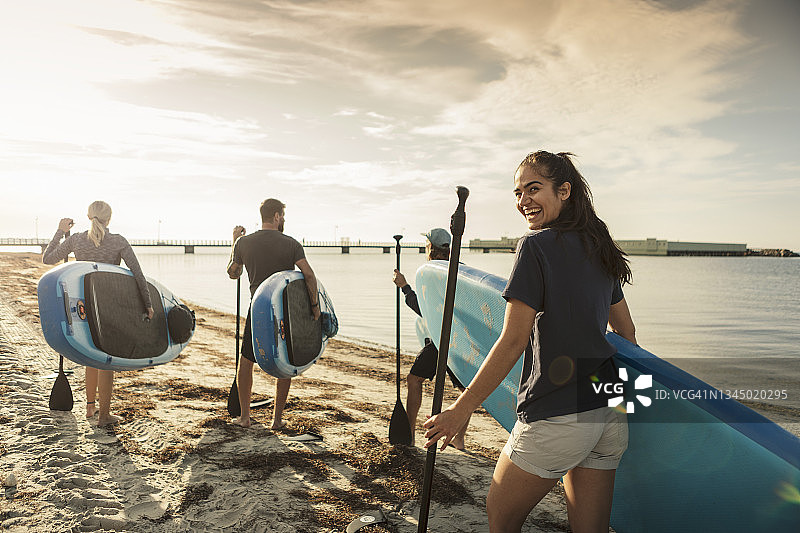 快乐的女人在海滩上与朋友们一起拿着桨板走路图片素材