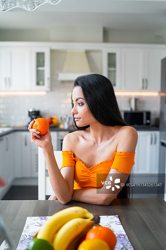 年轻漂亮的亚洲黑发女子坐在厨房的桌子旁，桌子上放着一面镜子，手里拿着一个橙子图片素材