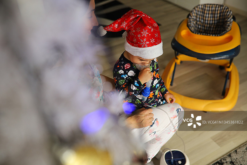 圣诞节的早晨，细心的母亲用雾化器为蹒跚学步的儿子吸入药物图片素材