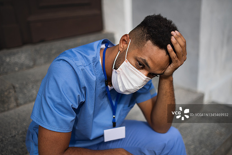 疲惫的男护工、男护士或戴着口罩的医护人员坐在室外诊所前的楼梯上图片素材