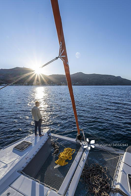 年轻男子站在双体船上看日落，提洛斯岛航海之旅，希腊多德卡尼斯图片素材