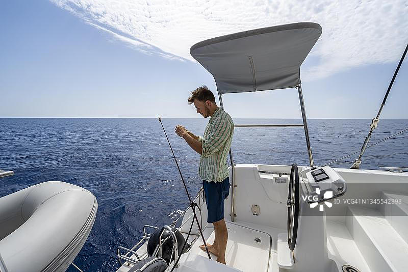 年轻男子在船上钓鱼，航行在希腊多德卡尼斯群岛的双体船上图片素材