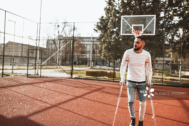 拄着拐杖腿上绑着绷带在篮球场上行走以恢复健康的男人图片素材