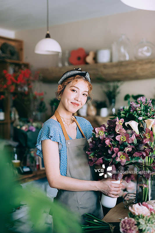 自信的年轻亚洲女花店主，小花店老板的肖像。站在她的商店里，拿着一束花。她面带微笑，看着镜头。享受和花在一起的工作。小企业概念图片素材