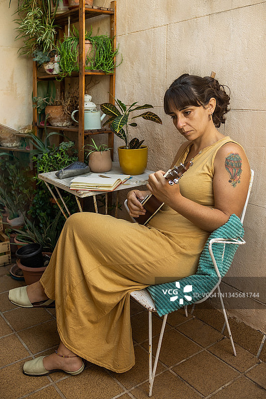 年轻女子身穿赭色连衣裙在阳台上弹奏尤克里里琴的侧面照图片素材