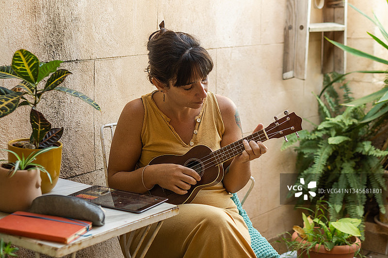 身穿赭色连衣裙的妇女在露台上练习尤克里里，周围环绕着植物和花盆图片素材