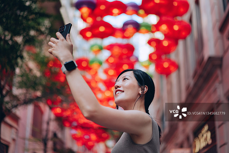 笑容甜美的年轻亚裔女子在挂满中国传统红灯笼的城市街道前，用手机自拍。图片素材