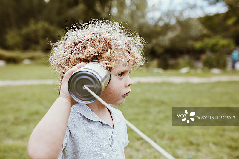 在草地上用罐头电话听筒通话的可爱金发男孩图片素材