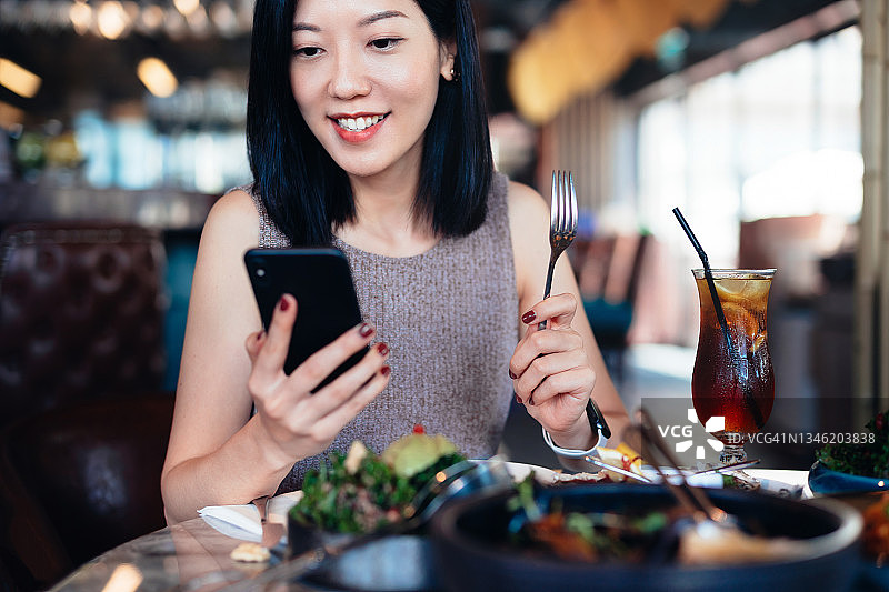 微笑的亚洲年轻女子在餐厅里看着她的智能手机，餐桌上新鲜美味的饭菜。外出就餐的生活方式。生活方式与科技图片素材