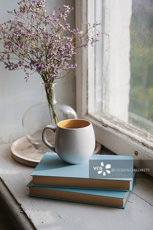 家居室内细节：杯子、书籍和带有紫色花朵的复古玻璃花瓶，摆放在木制窗台上图片素材