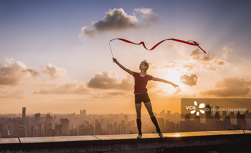 在美丽的日落和现代城市背景下手持彩带跳舞的快乐年轻女子图片素材