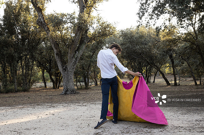 阳光明媚的日子里，优雅的男斗牛士在草地上练习，披着黄色和粉色的斗篷的背影图片素材