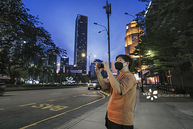 亚洲华人中年女性游客戴着口罩在城市霓虹灯下用手机拍照图片素材