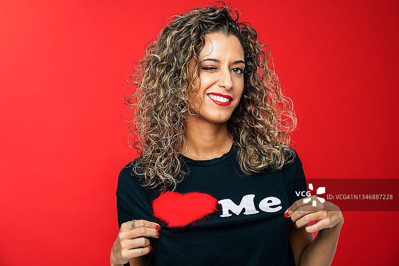 自信的年轻女子，有着卷曲的金发，对着镜头眨眼。她穿着牛仔裤和休闲黑色T恤。在鲜艳的红色背景下的工作室肖像。图片素材