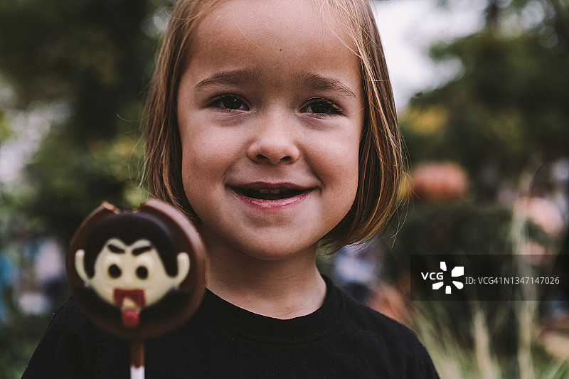 万圣节拿着吸血鬼图案巧克力棒棒糖在街上微笑的金发小女孩图片素材