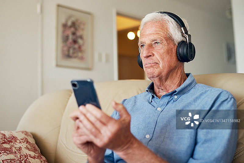 一位年长的男人在家中沙发上使用耳机和智能手机图片素材