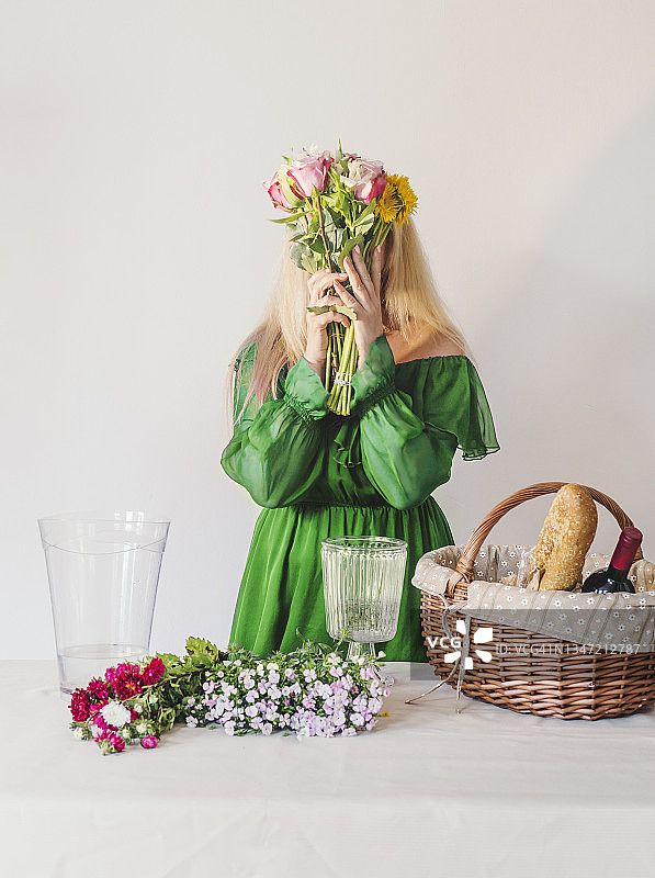 身穿绿裙的女人手捧鲜花花束，桌上有野餐篮、红酒、面包、鲜花和玻璃花瓶，白色墙壁背景图片素材
