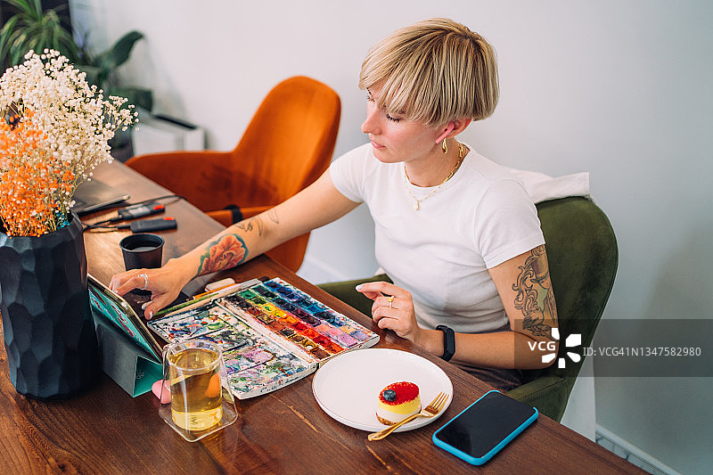 一位美丽的、真实的纹身金发短发女人正在观看课程，准备开始绘画。她坐在咖啡馆里喝茶和吃甜点。随时随地发挥创意爱好图片素材