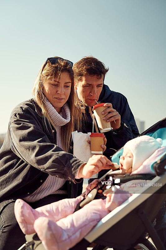 一对夫妇坐在长椅上休息，婴儿车里放着婴儿，一起喝咖啡图片素材