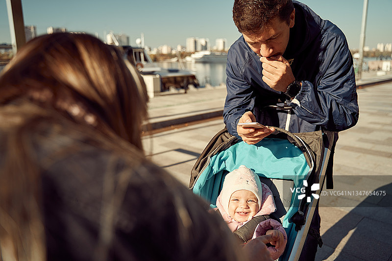 父亲看手机，年轻母亲在城市街道上用婴儿车和宝宝玩耍图片素材
