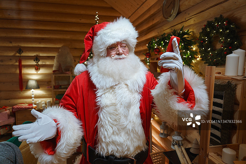 圣诞老人站在装饰着花环的圣诞树前，在他的木屋里通过智能手机上的视频电话与孩子们交流图片素材