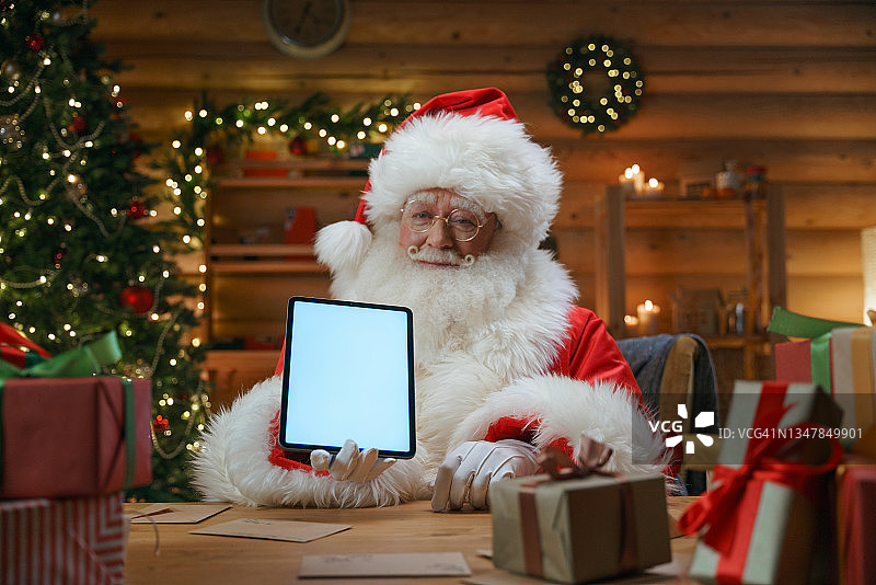 圣诞老人坐在小屋里，有圣诞装饰，在相机中展示数字平板电脑图片素材