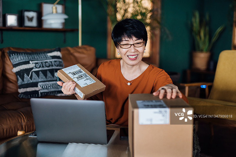 快乐的亚洲妇女在家里用笔记本电脑在线购物，收到一些包裹的送货服务，不能等待打开箱子的购买。网上购物，网上银行，愉快的客户购物体验图片素材