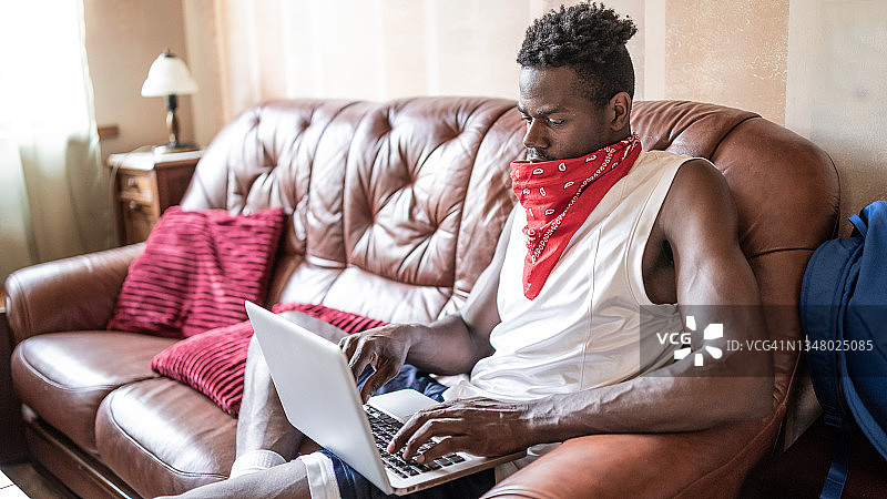 非洲男人在家用笔记本电脑在沙发上休息，看着屏幕，享受着慵懒的自由时光。生活方式概念。居家办公。图片素材