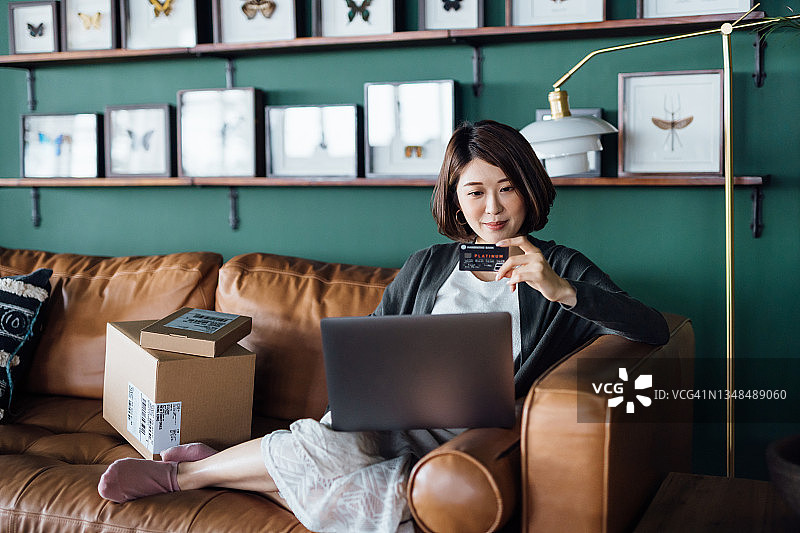 年轻亚裔女性在家中沙发上放松，使用笔记本电脑和信用卡在线购物，旁边堆放着已送达的包裹。图片素材