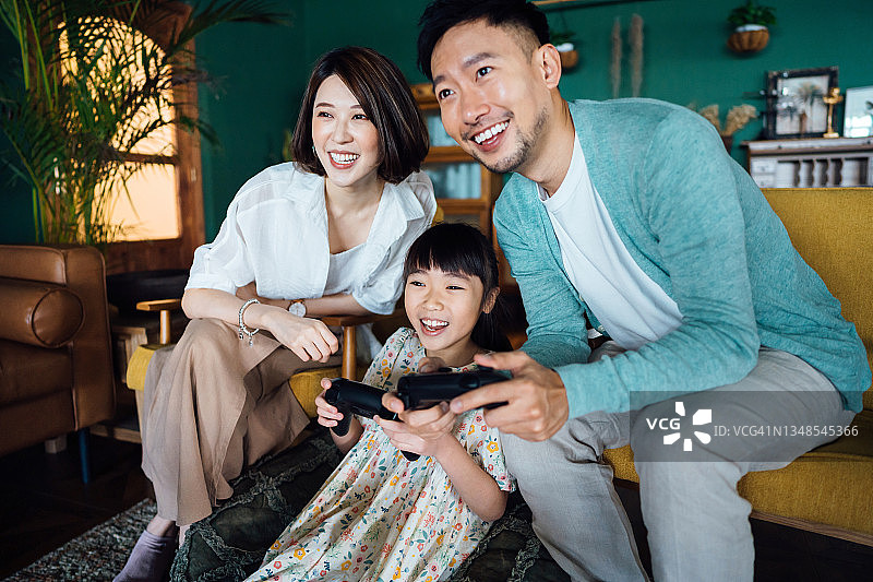 快乐的年轻亚洲家庭和女儿在客厅的沙发上玩电子游戏，共度美好时光图片素材