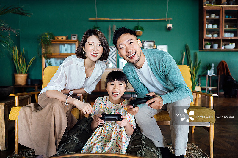 快乐的年轻亚洲家庭和女儿在客厅的沙发上玩电子游戏，共度美好时光图片素材