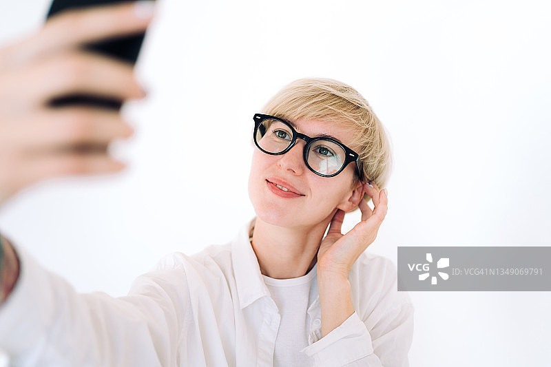 戴眼镜的金色短发年轻女人用智能手机自拍。她穿着白色休闲中性服装。性别中立概念图片素材