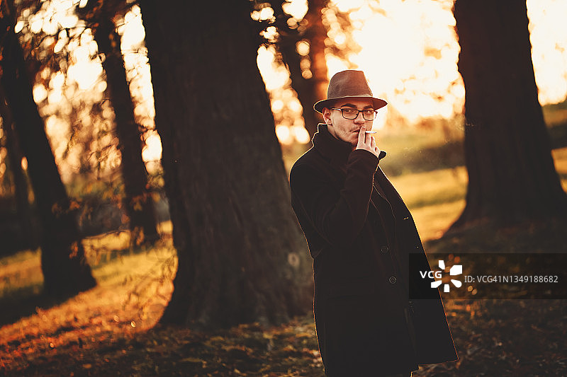 在秋天的公园里吸烟的戴帽子英俊男人图片素材