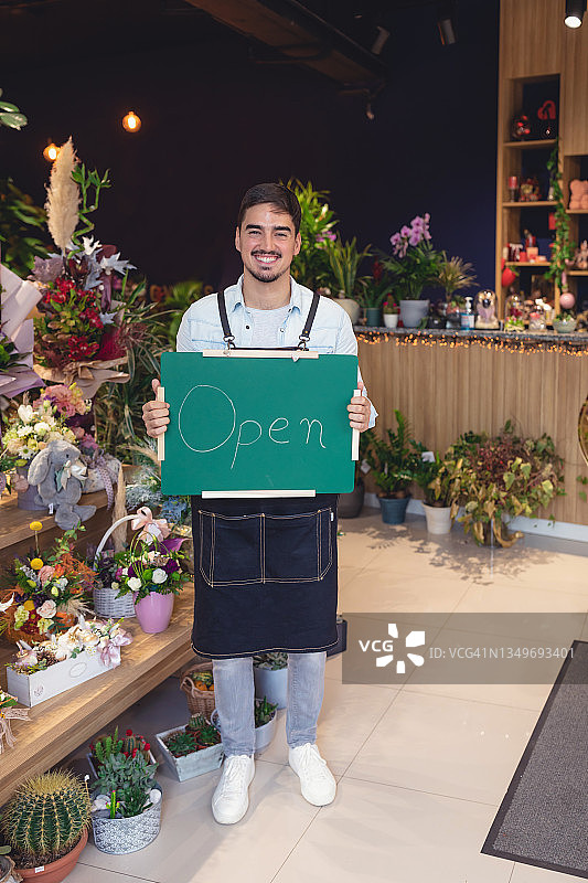 花店年轻的白人男店主手持Open标牌，对着镜头微笑特写。快乐的帅哥花店员工戴着口罩站在室内。商业概念图片素材