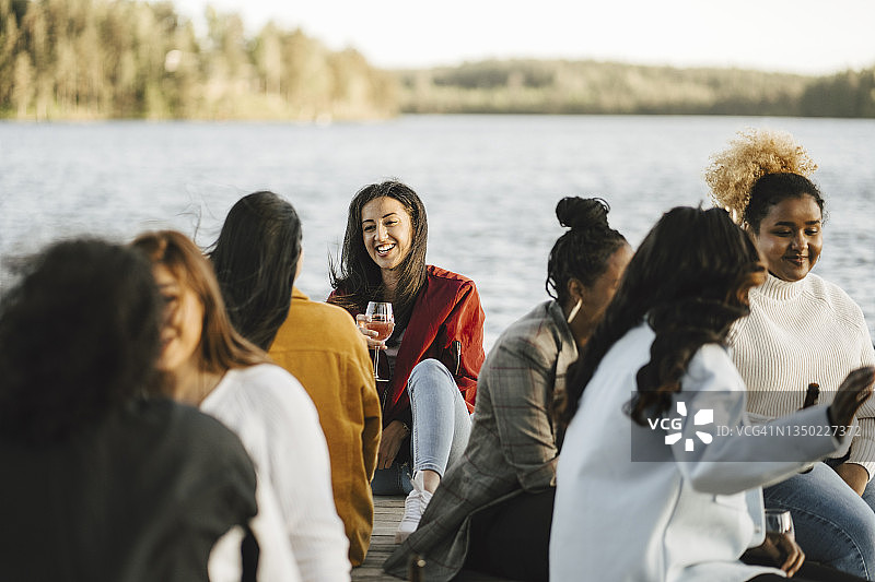 微笑的多种族女性朋友坐在码头上，背景是湖图片素材