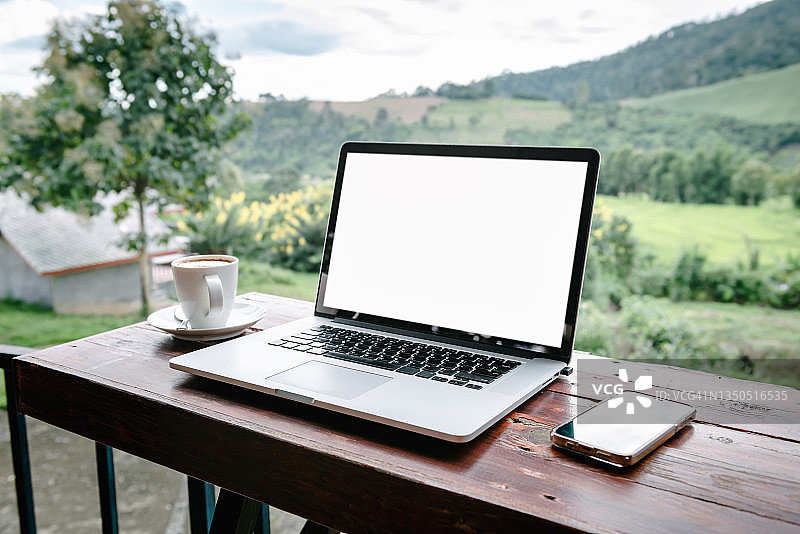 咖啡馆内山景背景下，桌子上带空白白色屏幕和手机的笔记本电脑图片素材