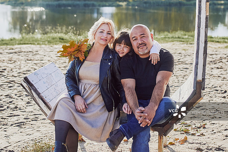 在阳光明媚的秋日，一个多种族家庭坐在河边的木凳上图片素材