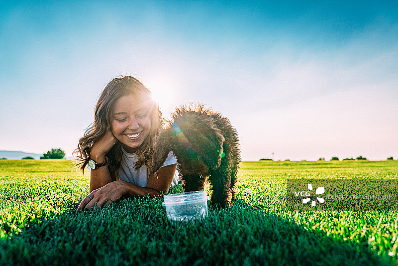 年轻女子坐着观看她的宠物“绵羊杜德尔犬”从可重复使用的塑料容器中喝水图片素材