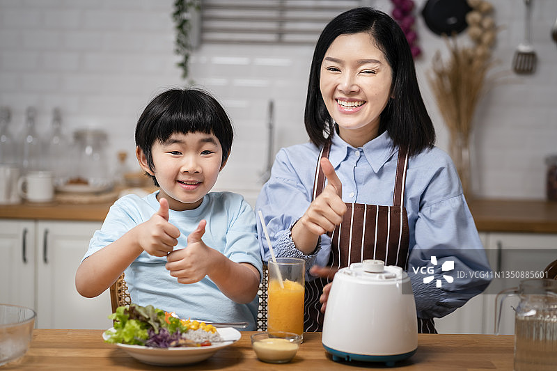亚洲母亲和儿子在厨房里做着大拇指手势，因为他们对自己的厨艺感到自信和自豪。图片素材