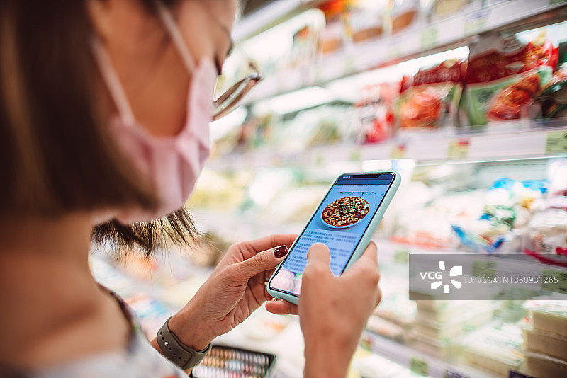 戴防护口罩的年轻女子在超市查看智能手机上的食谱并购买食材图片素材