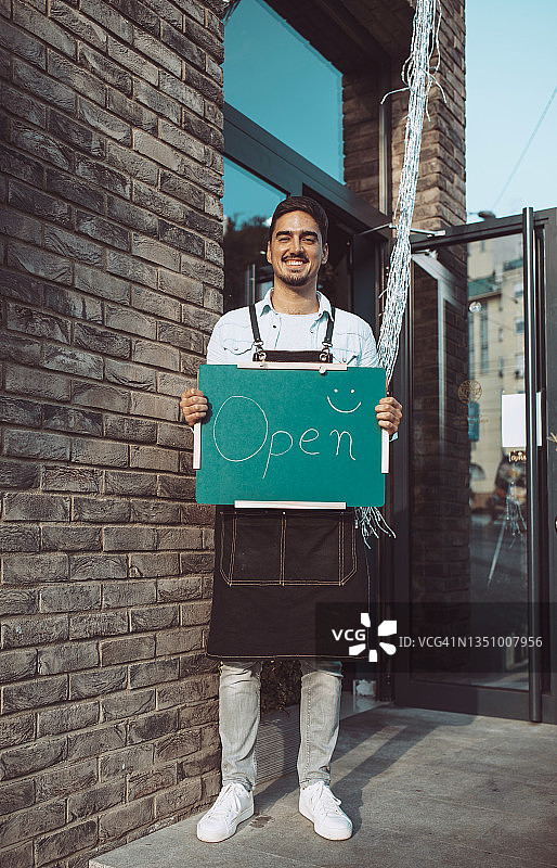 年轻的白人男性花店店主手持“营业中”标志，对着镜头微笑图片素材