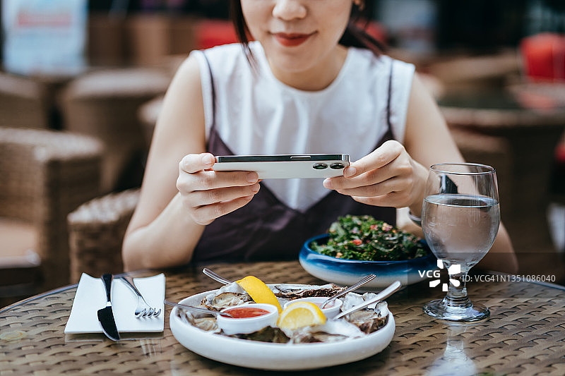 微笑的年轻亚洲女性在户外餐厅用餐前，用智能手机拍摄餐桌上新鲜美味食物的照片。外出就餐的生活方式。“相机先吃”文化图片素材