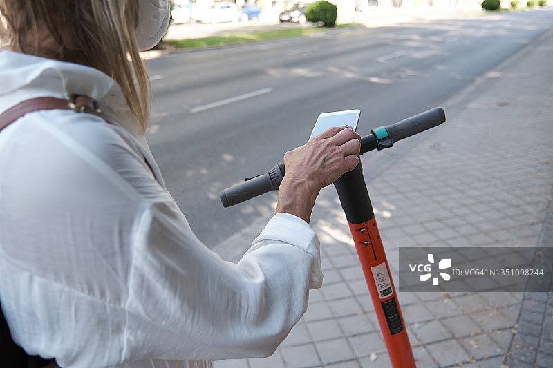 戴着防护口罩在城市里租用电动滑板车的匿名女人图片素材