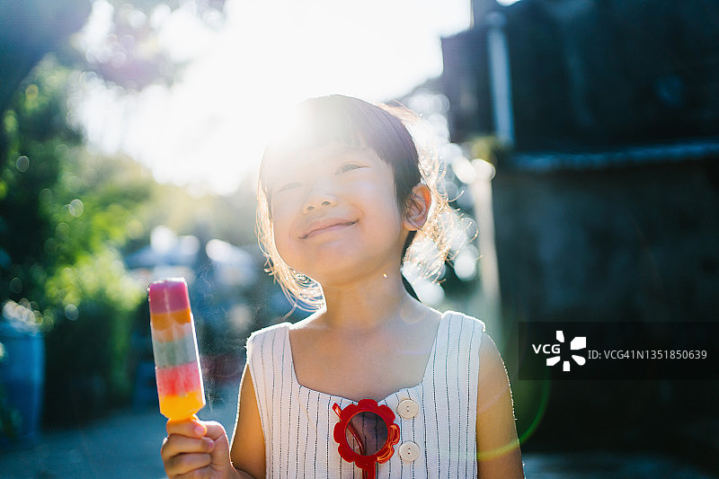 快乐的亚洲小女孩戴着红色太阳镜，在炎热的夏日户外吃着五彩缤纷的冰棒，站在有树木和天空的自然景色前图片素材
