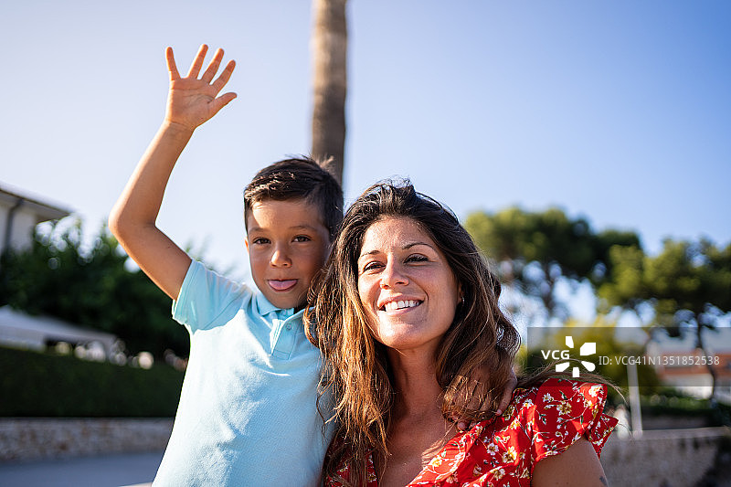 男孩在户外对着镜头挥手并做鬼脸，旁边站着一位女士图片素材